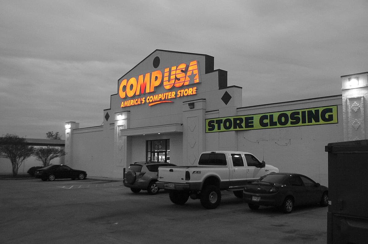 A CompUSA store with an illuminated orange sign and a yellow store closing banner, the rest of the photo in grey