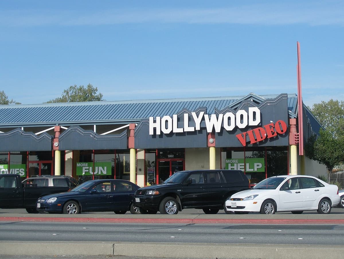 Hollywood Video storefront with white block letters and a red script word on a dark zigzag fascia, cars parked in front